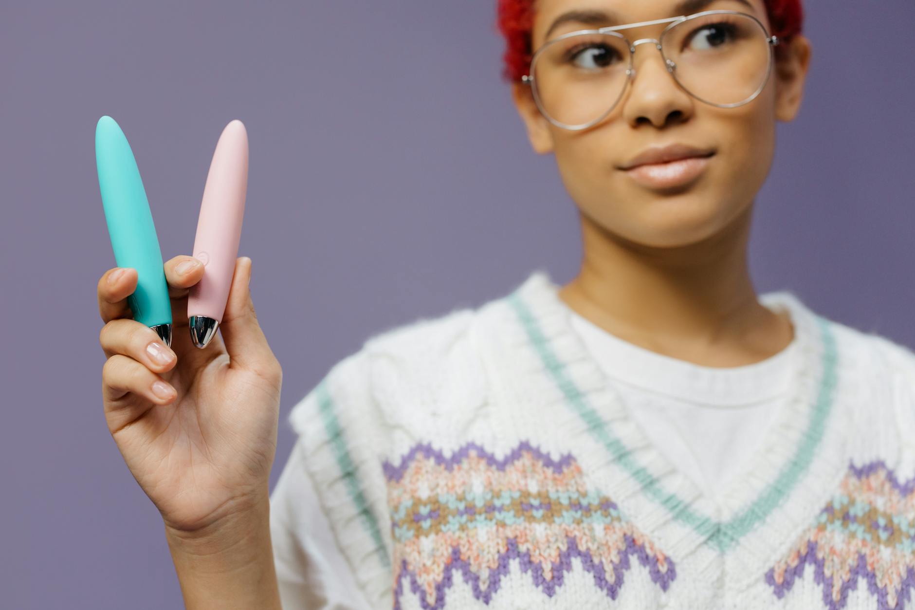 Woman holding two colorful vibrators against a purple background, representing pleasure and sexual wellness tools