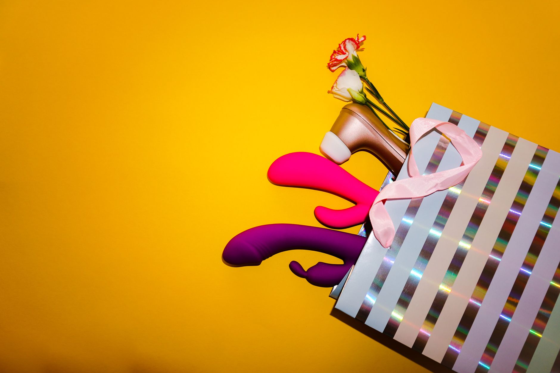 Colorful vibrators and flowers in a holographic gift bag on a bright yellow background